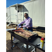 A man smiles while cooking sausage and bacon on a flat top grill. 