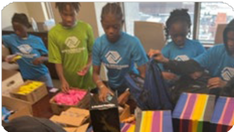 A few of the children from the Boys and Girls club stand at a table and fill backpacks with various items.