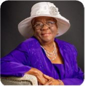 Janice Stanley sits wearing a large hat, with her arm resting on the back of a chair and smiling sightly. 