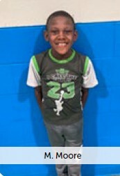 M. Moore. A little boy smiles standing in front of a brick wall.
