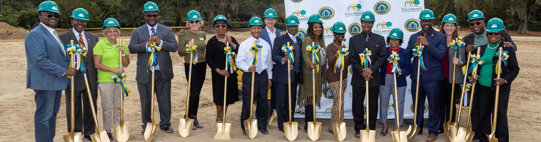 Group of people wearing green hard hats and holding gold shovels at groundbreaking event